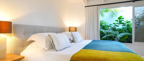 Una cama bien hecha con ropa de cama blanca y azul, una mesita de noche de madera y una ventana con vistas al verdor.