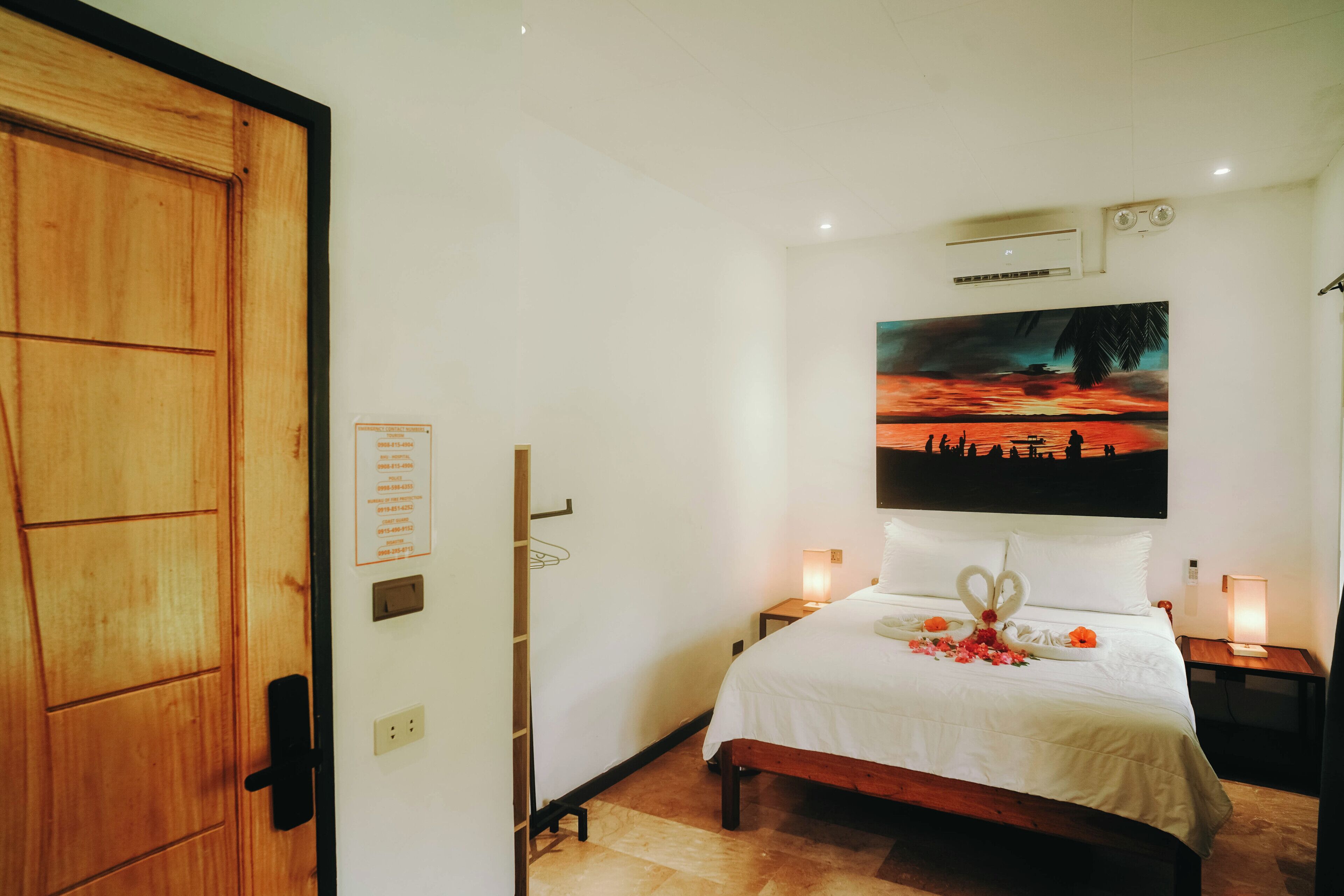 Habitación de hotel con una cama, un cuadro de un atardecer, una puerta de madera y un aire acondicionado instalado en la pared.
