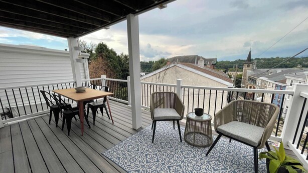 En overdækket terrasse med et bord og stole, et mønstret tæppe og udsigt over byen.