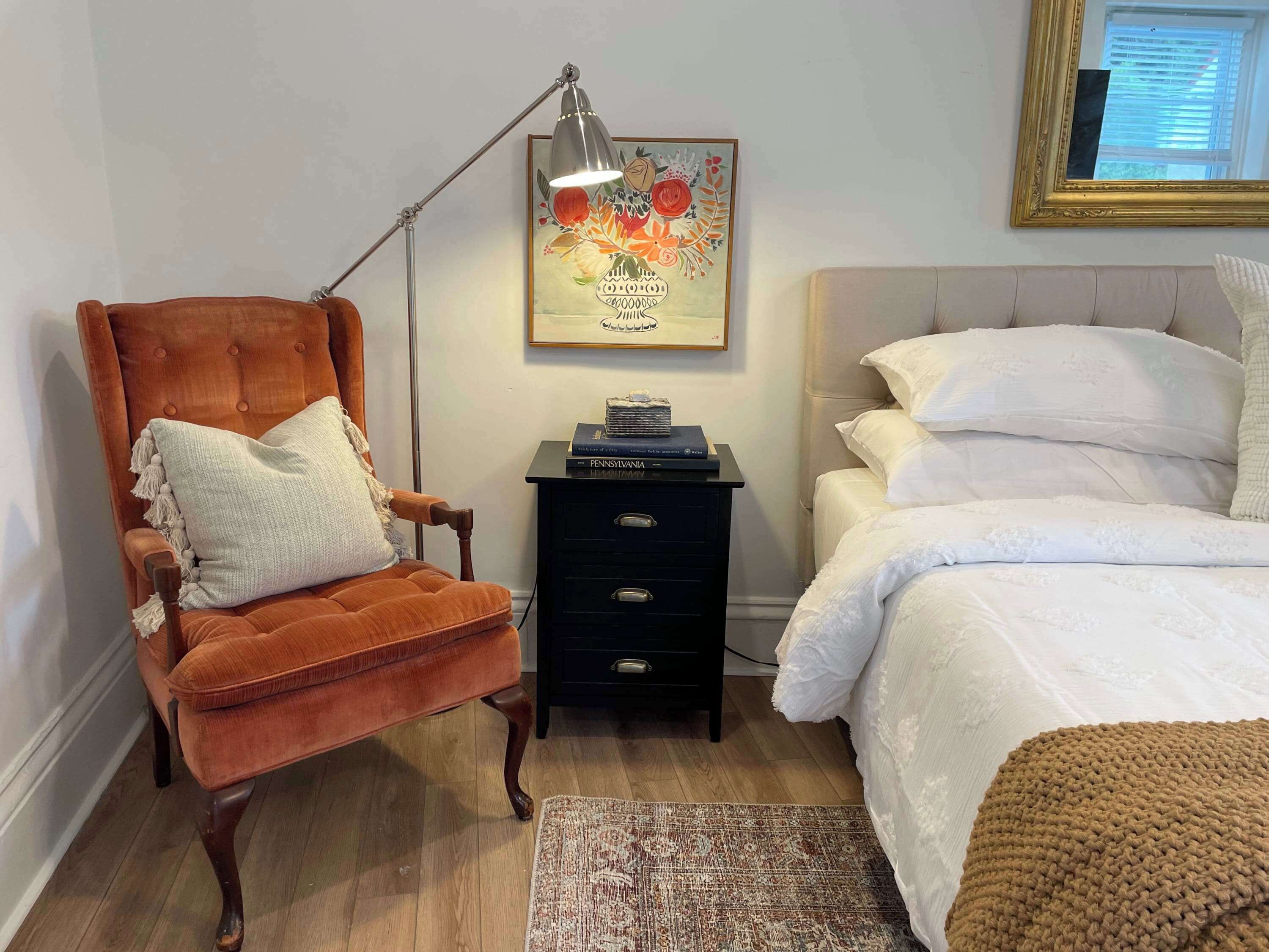 Une chambre avec un fauteuil marron, une table de chevet avec des livres, un lit avec une literie blanche et une lampe fixée au mur.