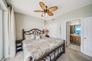 A bedroom with a bed, a ceiling fan, a nightstand, a dresser, a bathroom with a sink and mirror, and a door leading to another room. - Ocean Reef 1108 Beachfront, Heated Indoor & Outdoor Pools, Hot Tub, FREE Golf (Panama City Beach)