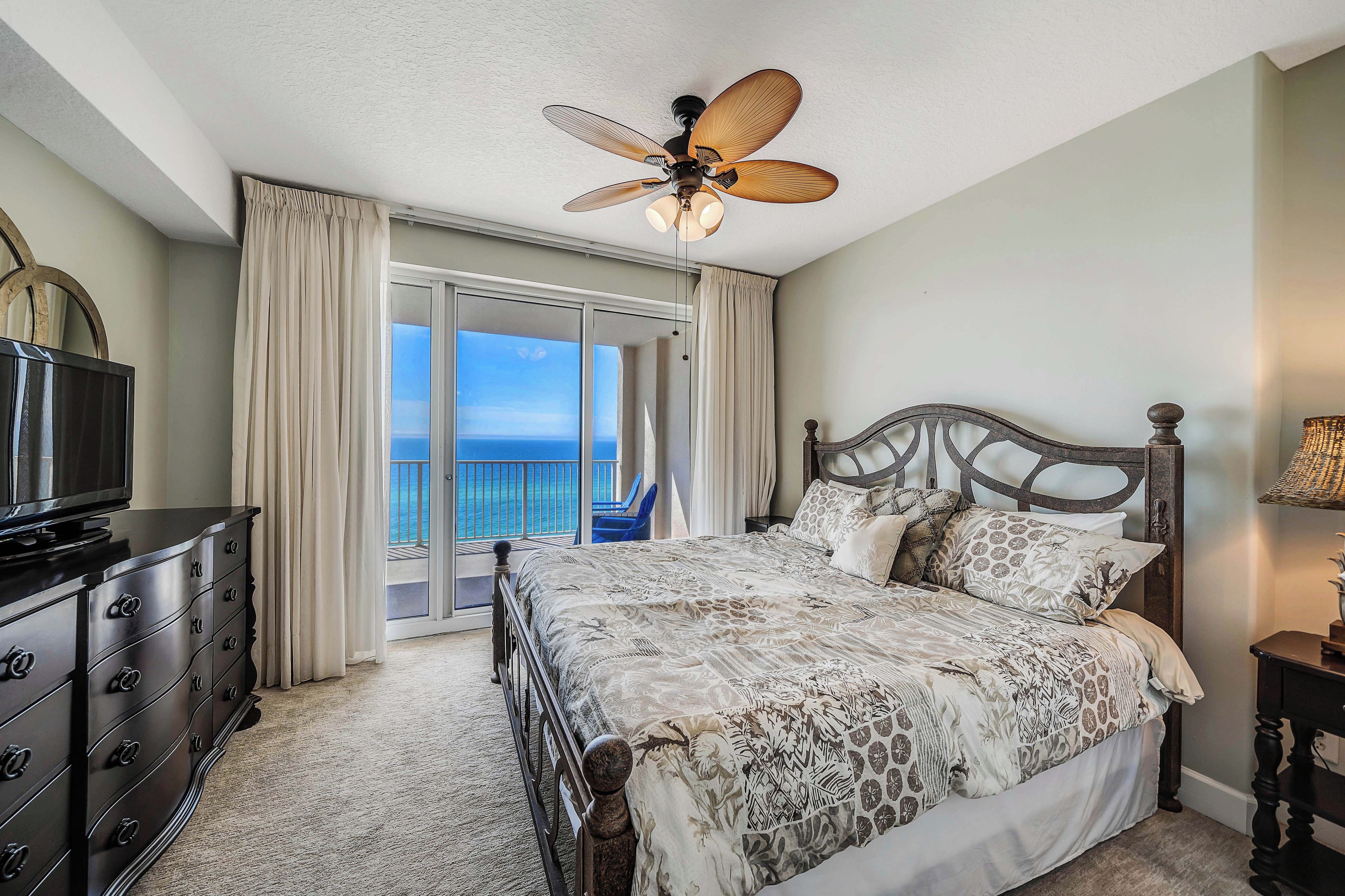 A bedroom with a bed, a television, a ceiling fan, and a view of the ocean through a sliding door.
