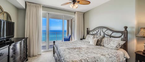 A bedroom with a bed, a television, a ceiling fan, and a view of the ocean through a sliding door.