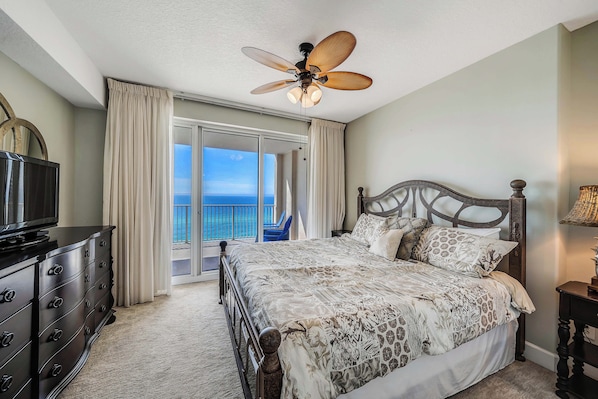 A bedroom with a bed, a television, a ceiling fan, and a view of the ocean through a sliding door.