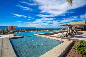 Zona de piscina con sillones de descanso y sombrillas, cielo despejado y vista a edificios y montañas distantes.
