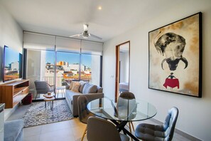 A modern living room with a glass table, sofa, and chairs, a large window with a city view, and a ceiling fan.