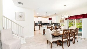 A spacious dining area with a large wooden table and chairs, a staircase with a white railing, and a view of the kitchen in the background. - ChampionsGate Home · Pool & Game Room Near Disney (Davenport)