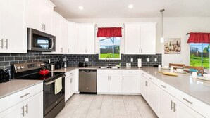 A modern kitchen with white cabinets, a black microwave, stainless steel appliances, and a red window valance. - ChampionsGate Home · Pool & Game Room Near Disney (Davenport)