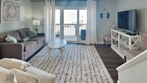 A living room with a sofa, a coffee table, a TV, and a balcony with curtains. - Dove's Landing | Serene Coastal Retreat + Access to Pool & More! (Surf City)