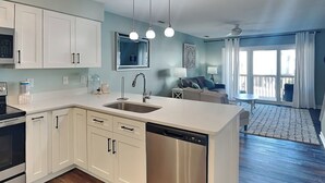 A modern kitchen with white cabinets, a stainless steel dishwasher, and a built-in sink. There is a dining area with a table and chairs, and a large window providing natural light. - Dove's Landing | Serene Coastal Retreat + Access to Pool & More! (Surf City)