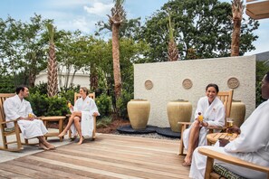A group of people in white robes seated on wooden chairs on a wooden deck, with large decorative vases and a textured wall in the background.
