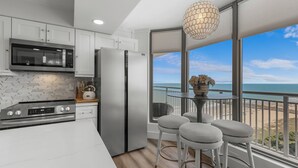 Cozinha moderna com eletrodomésticos de aço inoxidável, bancada branca e vista para a praia através de grandes janelas.