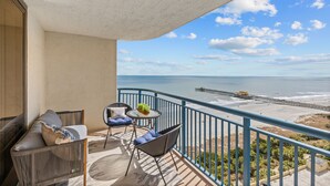 Uma varanda com vista para a praia e o cais, equipada com um sofá, cadeiras e uma mesa pequena.