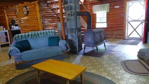 A cozy cabin interior with a wooden log wall, a blue sofa, a yellow table, and a wood-burning stove.