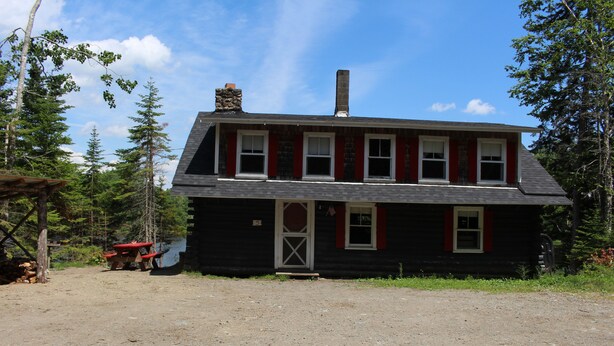 A two-story log cabin with a front porch, a chimney, and a small attached shed.