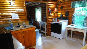 A rustic cabin interior with a wooden interior, a kitchen area including a stove, sink, and cabinets, and a window with a view of greenery outside.