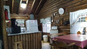 Ein rustikales Blockhaus-Interieur mit einer Holztheke, einem Kühlschrank, einer Kaffeemaschine und einem Esstisch mit Stühlen.