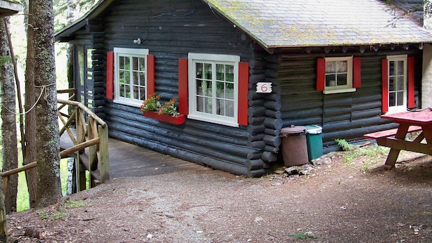Eine Blockhütte mit der Hausnummer 6, Holzfenstern mit roten Fensterläden und einem Picknicktisch vor der Tür.