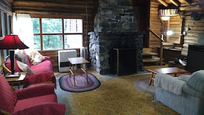 Ein gemütliches Blockhaus-Interieur mit Kamin aus Stein, Holzbalken, einem roten Sofa, einem grauen Sofa und einem kleinen Tisch.