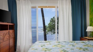 Une chambre à coucher avec un lit, un téléviseur posé sur un meuble en bois, une table de chevet avec une lampe, et une vue sur l’océan par une porte coulissante.