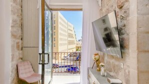 A room with a balcony, a TV mounted on the wall, a chair, and a console table with decorative items. - Luxury Apartment (Jerusalem)