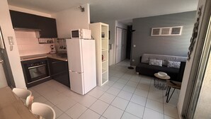 Une cuisine compacte équipée d’un réfrigérateur, d’un four et d’un four à micro-ondes. On y trouve une table à manger avec des chaises, ainsi qu’un canapé dans un coin salon dont le mur est de couleur grise.