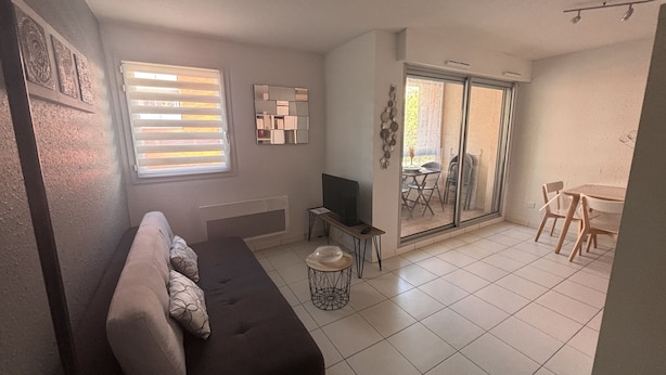 Un salon comprenant un canapé, un téléviseur, une petite table et une porte-fenêtre coulissante donnant sur un espace extérieur avec une table et des chaises.