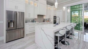 A modern kitchen with white cabinetry, stainless steel appliances, a marble island with bar stools, and pendant lighting.