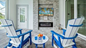 A covered outdoor seating area with two Adirondack chairs, a small table with drinks, and a fireplace with a painting above it.