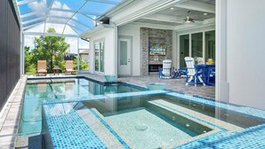 A modern indoor swimming pool with clear blue tiles, a glass ceiling, and a patio area with outdoor seating.