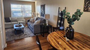Un salon comprenant un canapé, une table basse et une table à manger avec des chaises. Il y a une fenêtre avec des stores, une lampe et une plante sur la table.