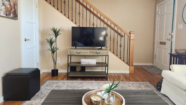 Un salon avec un escalier en bois, un téléviseur posé sur un meuble en bois, une plante en pot, un plateau avec des bougies et un canapé blanc.