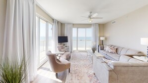 A spacious living room with a large window, a ceiling fan, a sofa, a TV, and a balcony with a view of the ocean. - HUGE Corner Unit Free Beach Chairs! Free round of Golf & Activities Daily! (Panama City Beach)
