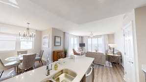 A modern kitchen with a central island, dining area with chairs, a living room with a sofa and TV, and large windows with curtains. - HUGE Corner Unit Free Beach Chairs! Free round of Golf & Activities Daily! (Panama City Beach)