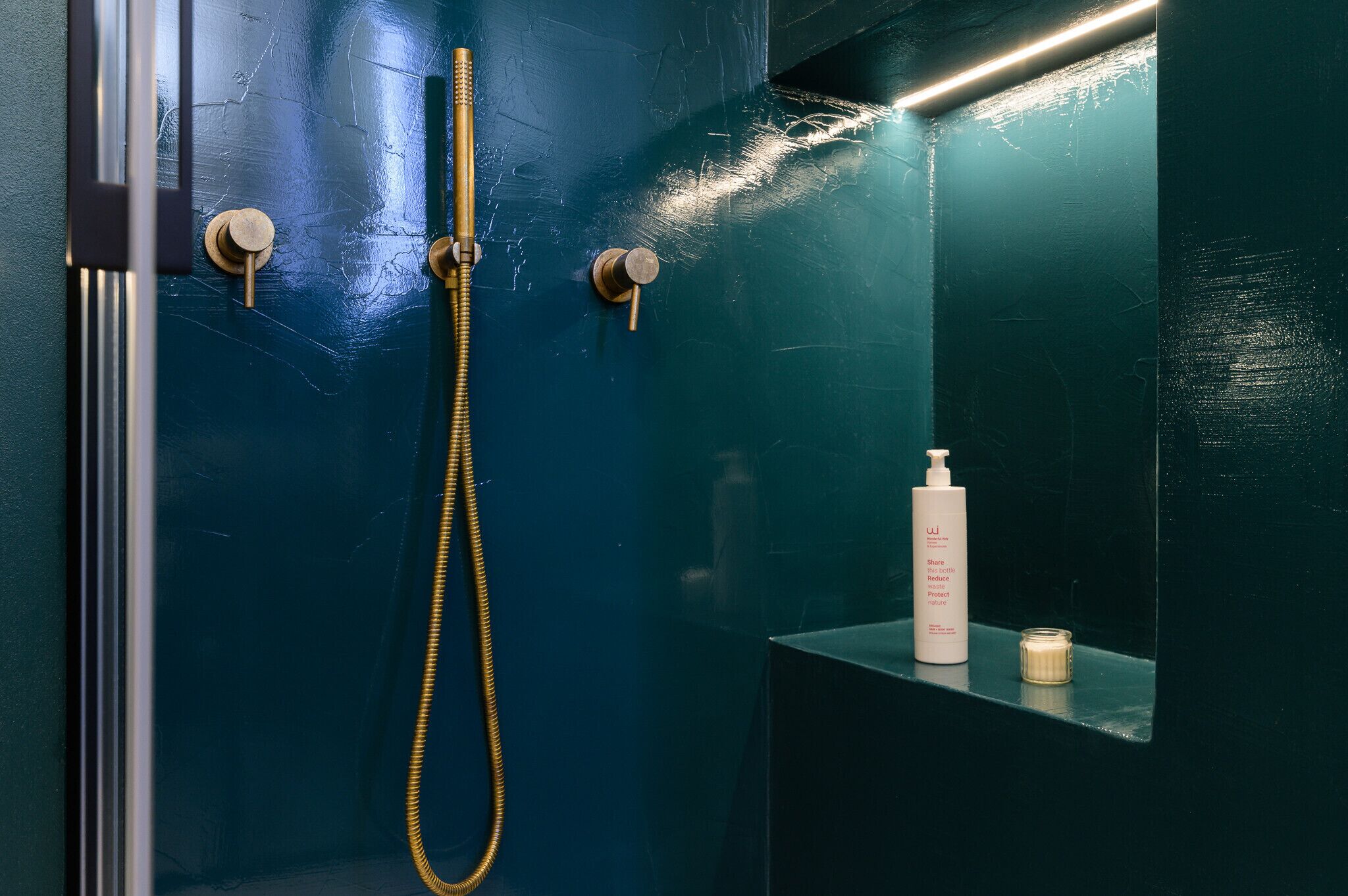 Une salle de bain avec un mur bleu foncé, une pomme de douche dorée et une étagère sur laquelle se trouvent une bouteille et un pot.