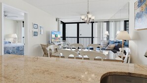 A modern living room with a dining area, featuring a chandelier, a sofa set, and a large window with curtains. - Sea Watch 1214 by Seaside Vacations & Sales (Ocean City)