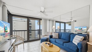 A modern living room with a blue sofa, a yellow coffee table, and a TV on a stand. There is a balcony with a view of buildings and a ceiling fan. - Sea Watch 1214 by Seaside Vacations & Sales (Ocean City)