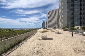 Ein Strand mit Liegestühlen und Sonnenschirmen, einem Zaun und hohen Gebäuden im Hintergrund.