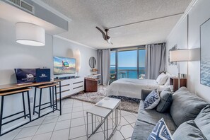 A modern hotel room with a bed, a desk, a TV, and a view of the sea.