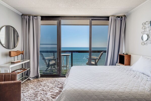 A bedroom with a large bed, a view of the sea, a balcony with chairs, and a round mirror on the wall.