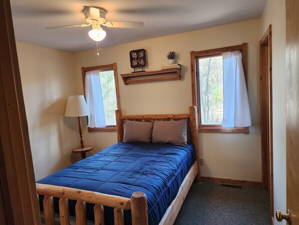 Ein Schlafzimmer mit einem hölzernen Bett, zwei Vorhängen an den Fenstern, einem Deckenventilator und einem kleinen Regal mit Büchern und einer Pflanze.