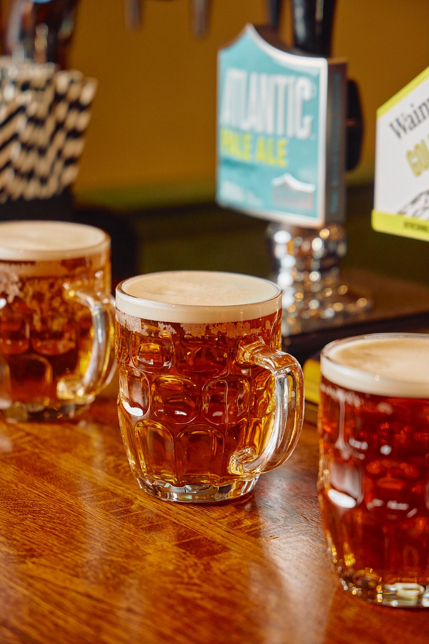 Drie bierglazen op een houten toog met een biertappuntje, gelabeld 'Atlantic Pale Ale'.