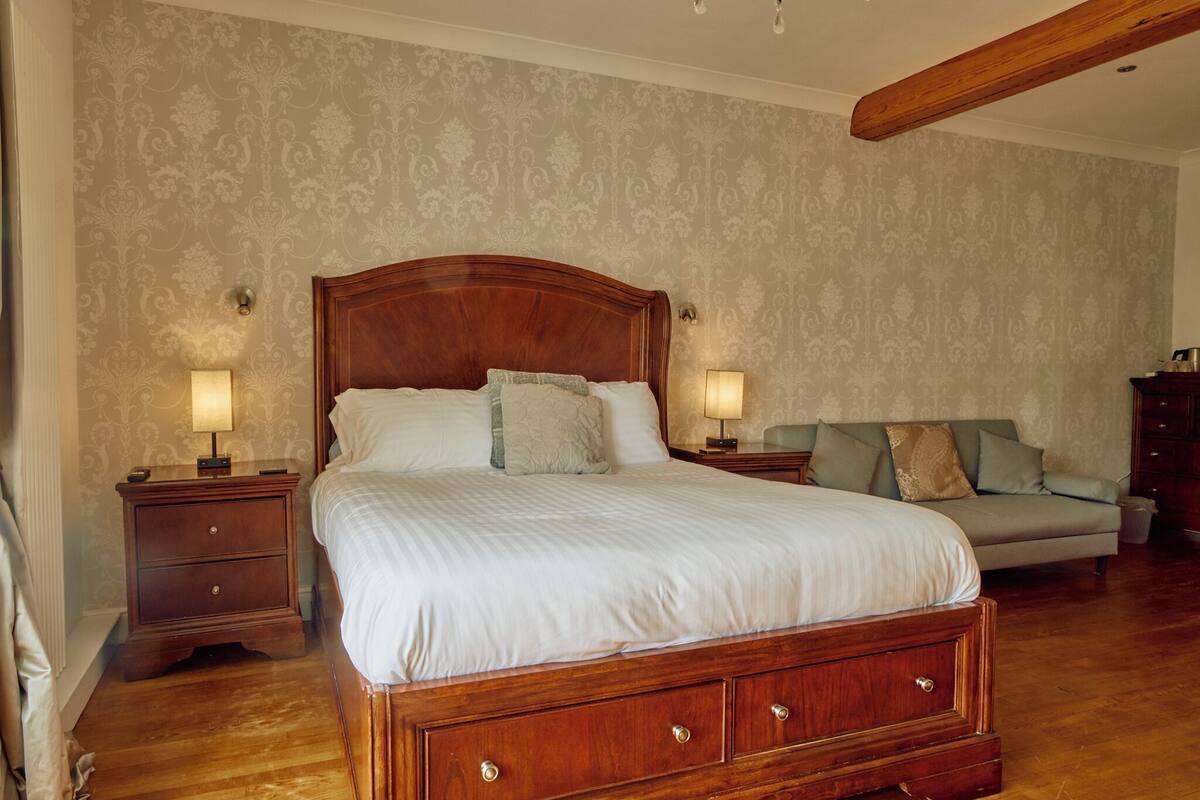 A bedroom with a large wooden bed, two bedside tables with lamps, a grey sofa, and patterned wallpaper.