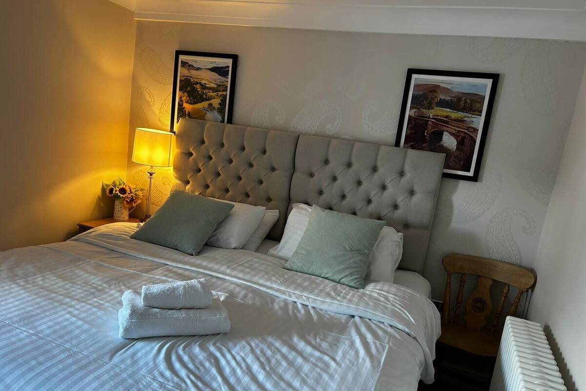 A neatly made bed with a tufted headboard, two framed pictures on the wall, a bedside lamp, and a small wooden nightstand.
