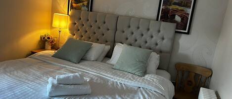 Una cama bien tendida con un cabecero capitonado, dos cuadros enmarcados en la pared, una lámpara de noche y una mesita de noche de madera.