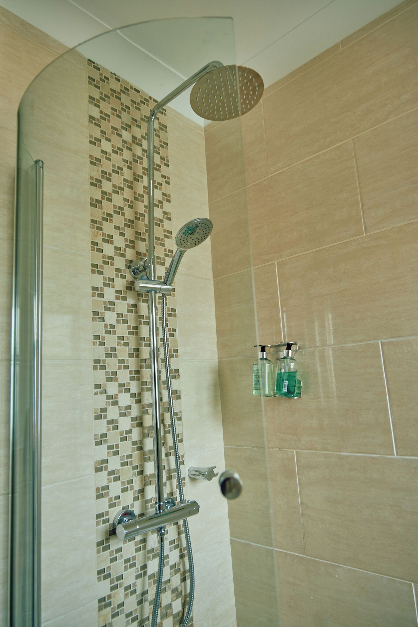 Una ducha con cabezal de ducha de lluvia, un cabezal de ducha manual y un accesorio de ducha manual. Hay dos botellas de champú y acondicionador en la pared.