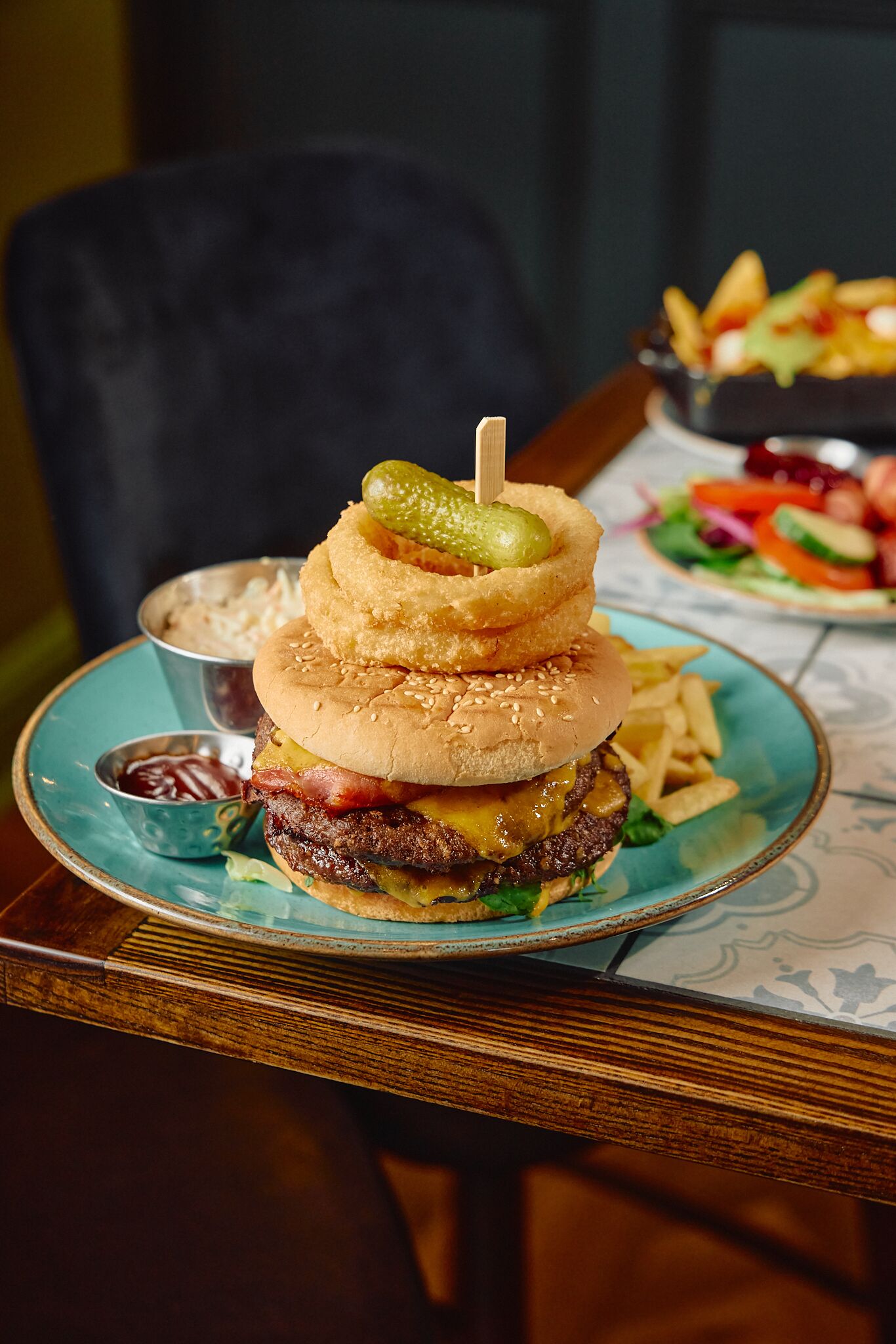 A burger with a pickle on top, served on a blue plate with fries and a small bowl of ketchup.