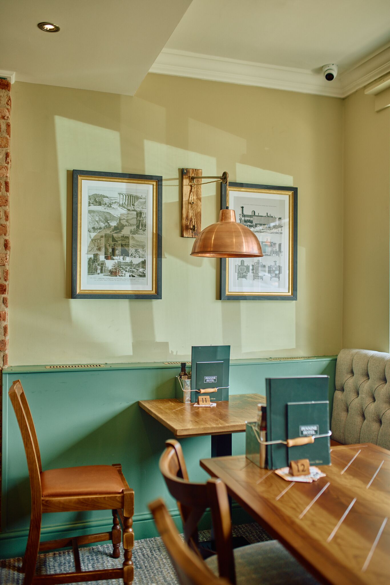 A dining area with a wooden table, chairs, and a green cushioned bench. There are framed pictures on the wall and a pendant light above the table.