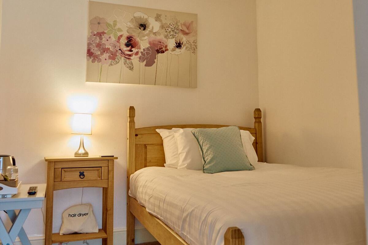 A bedroom with a wooden bed, a nightstand with a lamp, a wall-mounted floral artwork, and a small chair with a bag labeled "hair dryer".