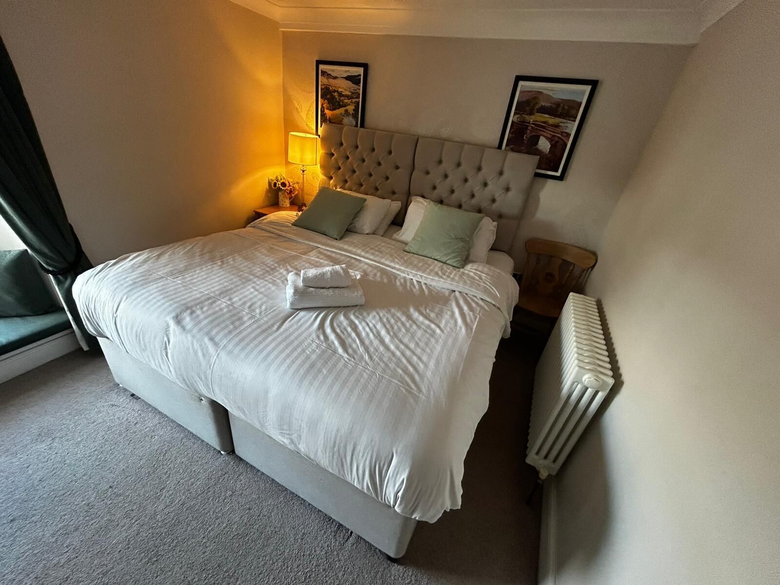 A neatly made bed with a tufted headboard, a bedside table with a lamp, and two framed pictures on the wall.